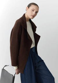Person wearing a brown coat and blue jeans holding a black bag against a white background
