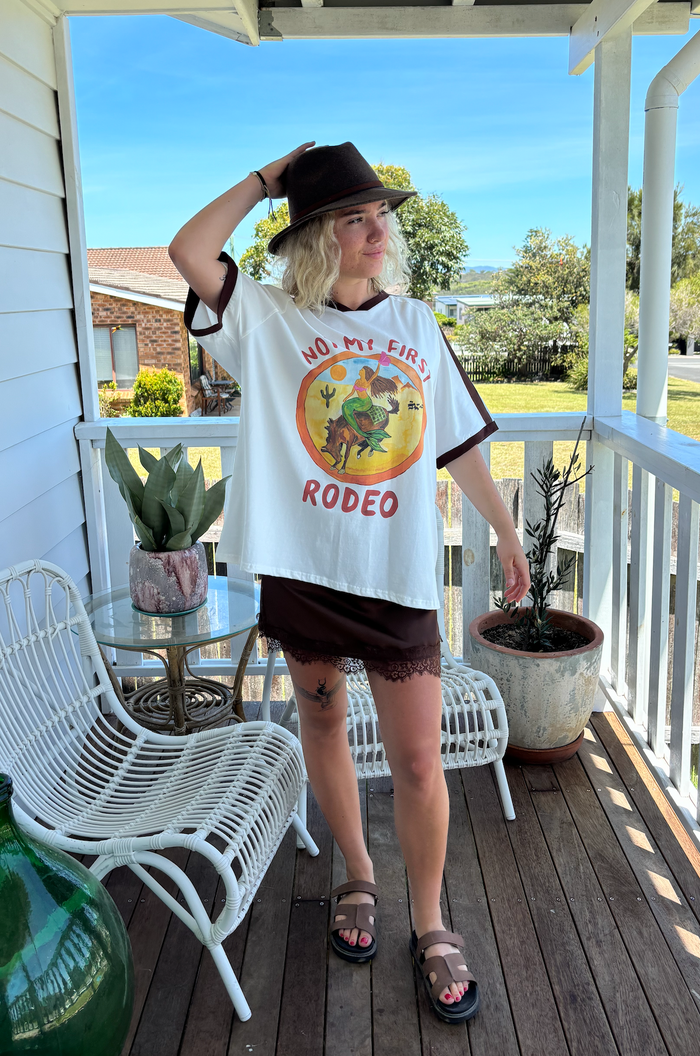 not my first rodeo tee by frankie is a white cotton tee with a mermaid riding a horse in a desert sccene with contrast sleeve stripes