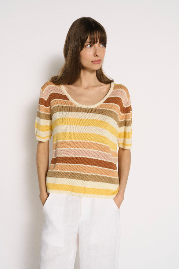 A woman wearing a short-sleeve knit top with horizontal stripes in yellow, orange, brown, and cream, paired with white trousers, standing against a plain white wall, by Little Lies