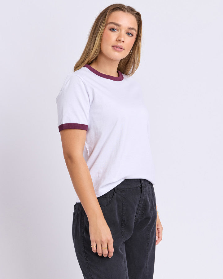 A woman modeling a white ringer t-shirt with dark purple trim on the crew neck and sleeve cuffs, paired with dark charcoal pants